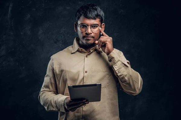 Självsäker indisk man i glasögon och skjortan håller en tablett dator och tittar på en kamera med en eftertänksam blick — Stockfoto