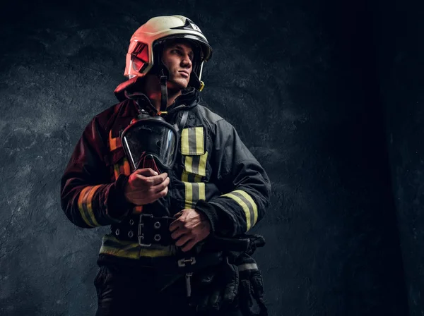 Bombeiro brutal em uniforme e capacete de segurança segurando uma máscara de oxigênio e olhando para o lado com um olhar confiante . — Fotografia de Stock