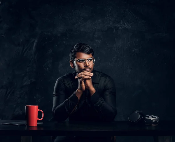 Indischer Student sitzt an einem Schreibtisch mit Laptop, das Kinn an die Hände gelehnt und seitwärts schauend. — Stockfoto