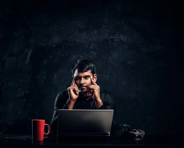 Indian student sitting at a desk with a laptop and look at the camera with a thoughtful look.