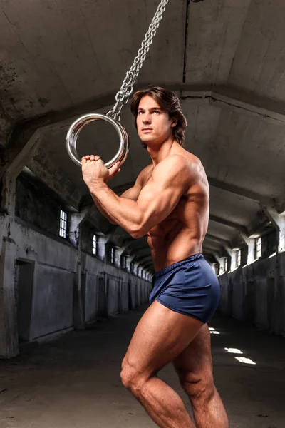 Un guapo gimnasta con cuerpo muscular posando con anillos de gimnasia en el abandonado edificio industrial —  Fotos de Stock