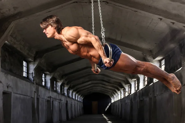 Ένας επαγγελματίας αθλητής με όμορφο μυώδες σώμα συρμών στο Γυμναστικά δαχτυλίδια σε εγκαταλελειμμένο βιομηχανικό κτίριο — Φωτογραφία Αρχείου