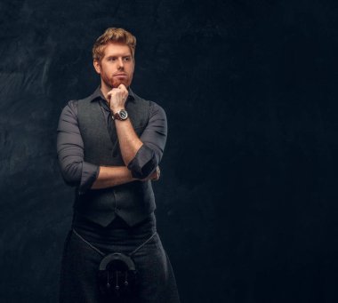 Redhead man dressed in an elegant vest with tie and kilt posing with thoughtful look in studio against a dark textured wall clipart