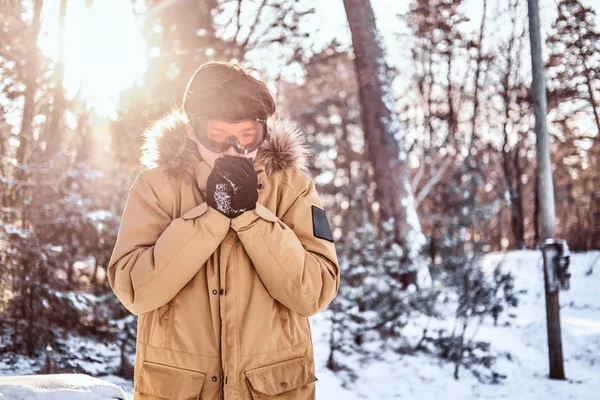 젊은 snowboarder 따뜻한 옷과 고글, 옷을 입고 눈 덮인 숲에 서 있는 그의 손을 따뜻하게합니다 — 스톡 사진