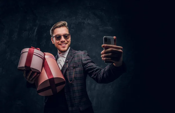 Um jovem vestido com um terno elegante segurando presentes e tirando selfie por smartphone em um estúdio escuro — Fotografia de Stock