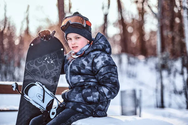 Ritratto di uno scolaretto carino vestito di tuta da neve seduto con uno snowboard su una panchina nella foresta invernale — Foto Stock