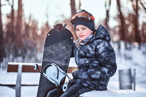 Retrato de un lindo colegial vestido con traje de nieve sentado con una tabla de snowboard en un banco en el bosque de invierno — Foto de Stock