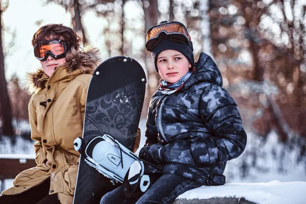 Due fratelli adolescenti vestiti con abiti caldi seduti su una panchina con uno snowboard in un parco — Foto Stock