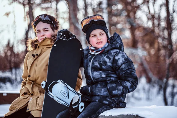 Dos hermanos adolescentes vestidos con ropa de abrigo sentados en un banco con una tabla de snowboard en un parque —  Fotos de Stock