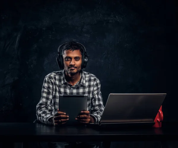 Μαθητή Ινδικό στα ακουστικά κρατώντας ένα δισκίο, να κάθεται σε ένα γραφείο και να βλέπουν μια φωτογραφική μηχανή. — Φωτογραφία Αρχείου