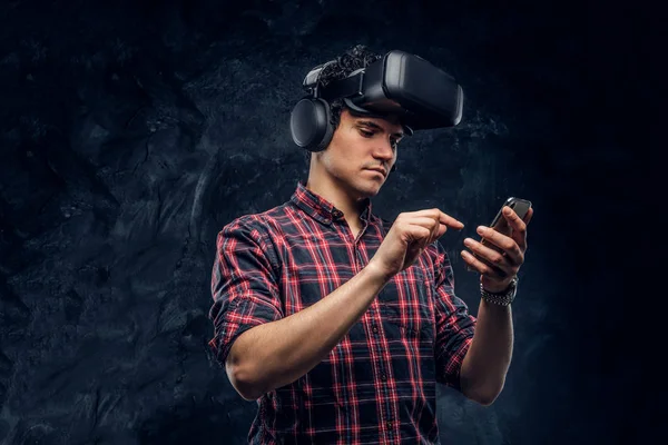 Хлопець у навушниках та гарнітурі VR, використовуючи смартфон у студії на фоні темної стіни — стокове фото