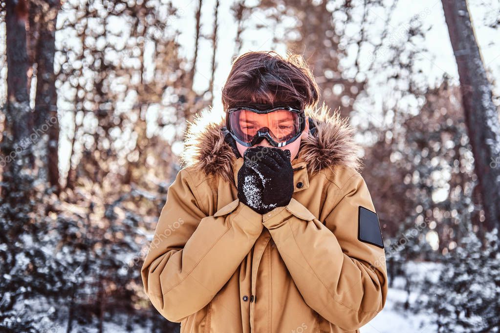 Young snowboarder dressed in warm clothes and goggles, warms his hands standing in the snowy forest