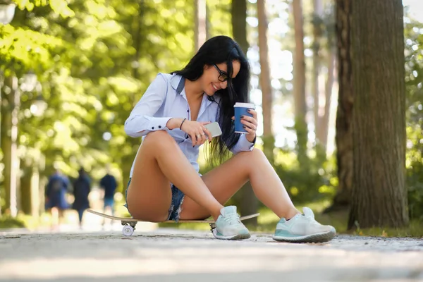 Красива дівчина в білій сорочці і шортах сидить на скейтборді в парку в літній сонячний день — стокове фото