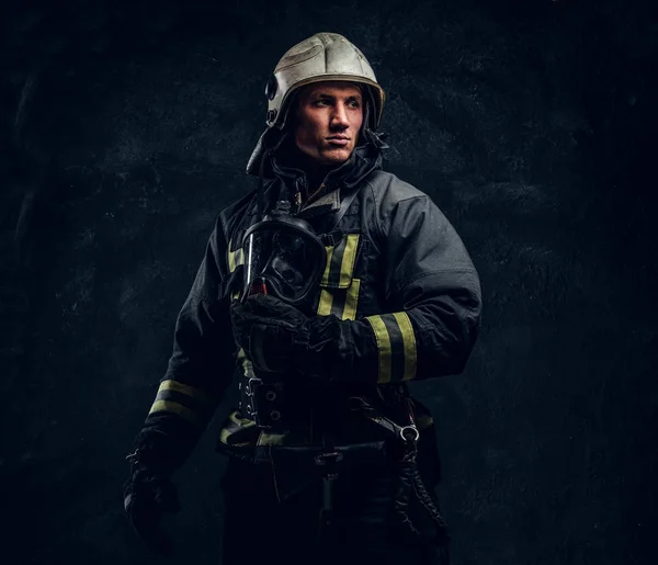 Bombeiro brutal em uniforme e capacete de segurança segurando uma máscara de oxigênio e olhando para o lado com um olhar confiante . — Fotografia de Stock