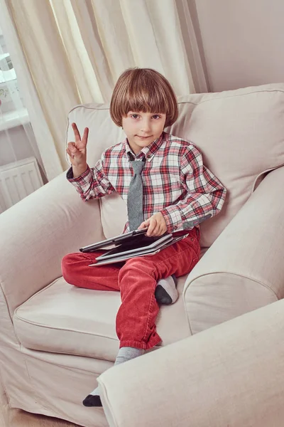 Estudante caucasiano vestindo uma camisa quadriculada com gravata mostrando sinal de paz e segurando um tablet enquanto estava sentado em um sofá em casa — Fotografia de Stock