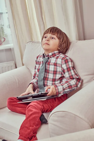 Skolpojke bär en rutig skjorta med slips håller en digital surfplatta medan du sitter på en soffa med en konstig blick hemma. — Stockfoto