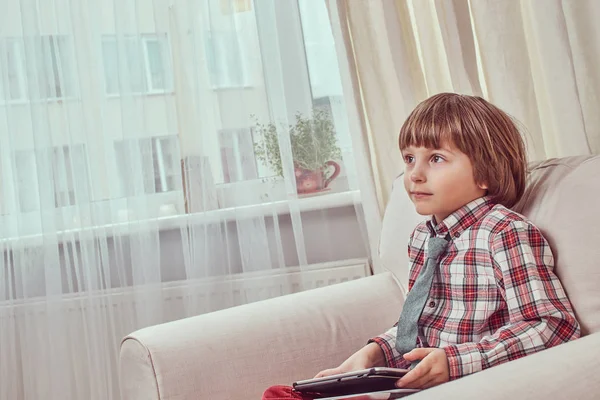 En stilig pojke som bär en rutig skjorta med slips håller en surfplatta medan du sitter på en soffa hemma — Stockfoto
