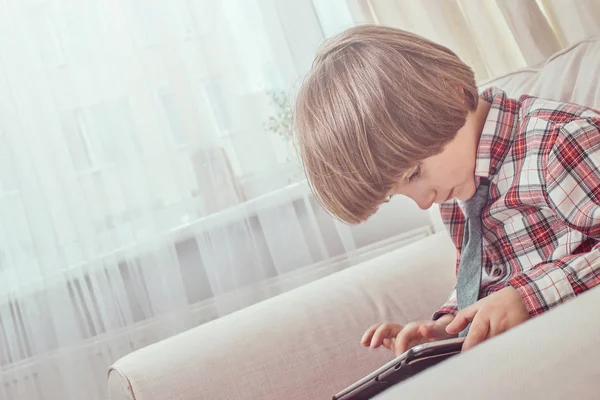 Kaukasiska skolpojke bär en rutig skjorta med slips med hjälp av en digital tablett som sitter på en soffa hemma — Stockfoto