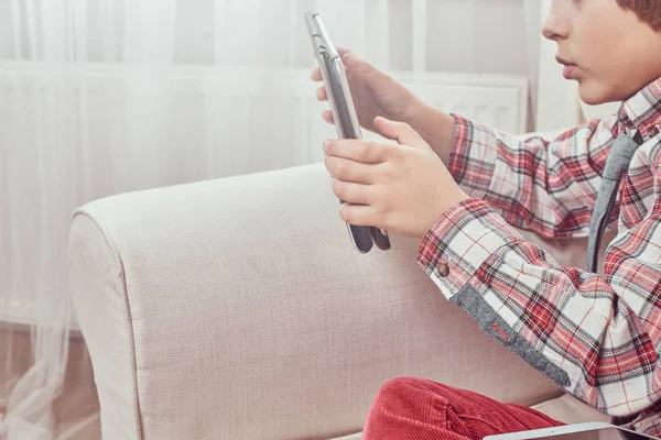 Sidovy av unga kaukasiska skolpojke bär en rutig skjorta med slips med hjälp av en digital tablett som sitter på en soffa hemma. — Stockfoto