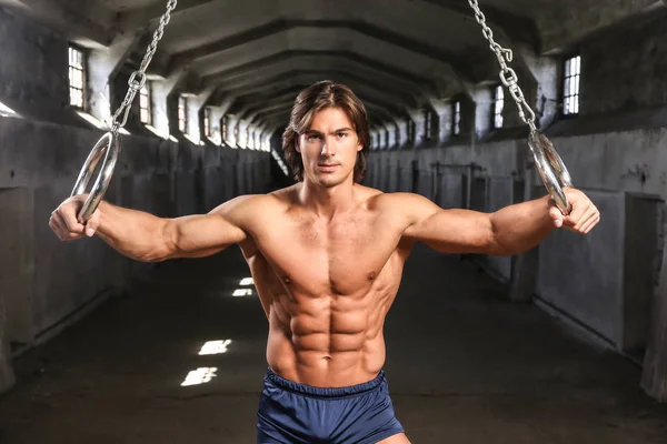 Un guapo gimnasta con cuerpo muscular posando con anillos de gimnasia en el abandonado edificio industrial — Foto de Stock