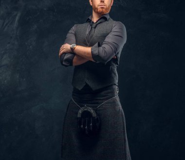 Redhead man dressed in an elegant vest with tie and kilt posing with his arms crossed in studio against a dark textured wall clipart