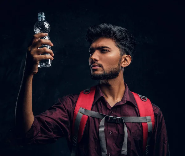 Indiase wandelaar met rugzak houden een fles water op zoek naar haar. Studio foto tegen een donkere getextureerde muur — Stockfoto