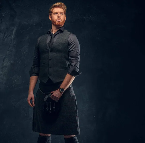 Redhead man dressed in an elegant vest with tie and kilt in studio against a dark textured wall