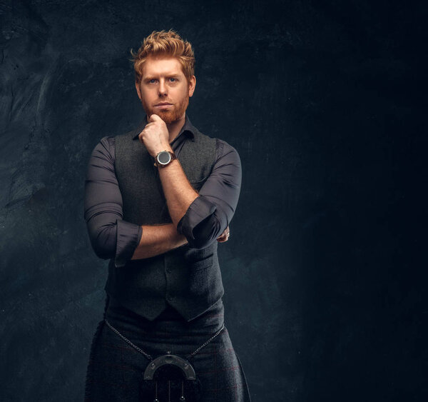 Redhead man dressed in an elegant vest with tie and kilt posing with thoughtful look in studio against a dark textured wall