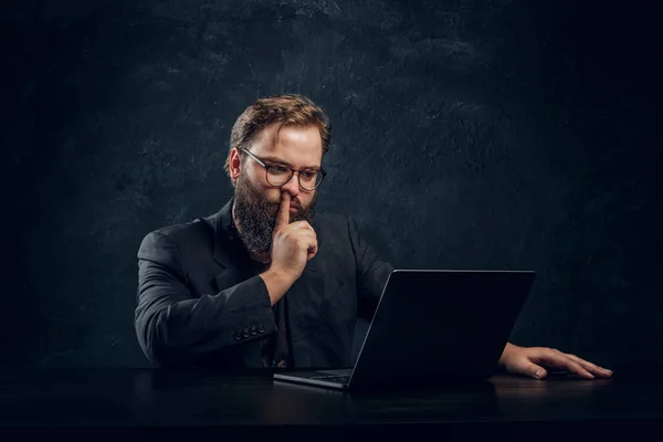 Retrato de un programador barbudo sentado en la mesa con portátil y con un dedo en la nariz . —  Fotos de Stock