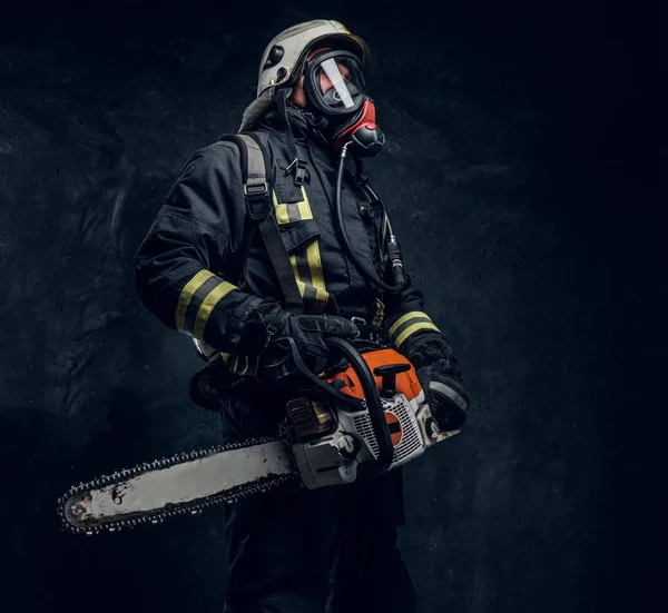 Retrato de un bombero con casco de seguridad y máscara de oxígeno sosteniendo una motosierra. Foto del estudio contra una pared de textura oscura — Foto de Stock