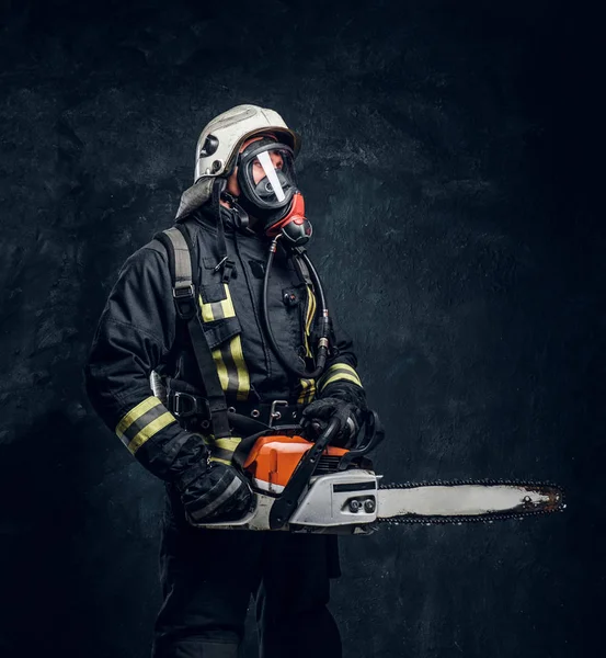 Retrato de um bombeiro em capacete de segurança e máscara de oxigênio segurando uma motosserra. Foto do estúdio contra uma parede de textura escura — Fotografia de Stock