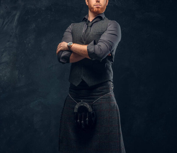 Redhead man dressed in an elegant vest with tie and kilt posing with his arms crossed in studio against a dark textured wall