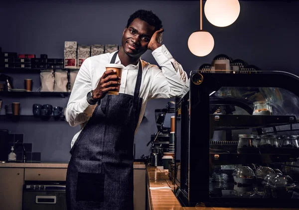 Cansado barista sosteniendo una taza con café mientras se apoya en un mostrador en una cafetería y mirando a una cámara con una mirada feliz — Foto de Stock