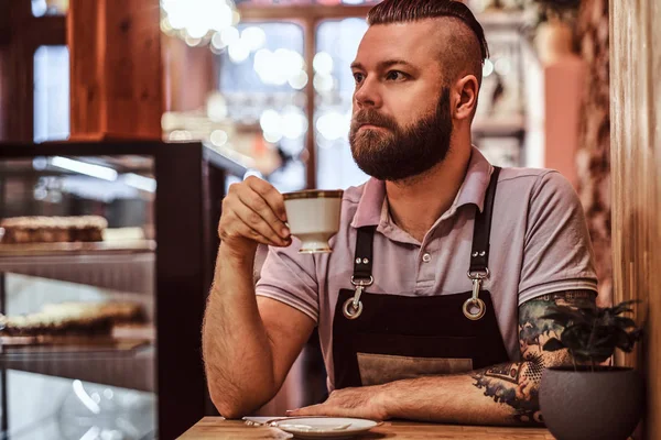 Bonito barista vestindo avental bebendo café durante a pausa para o almoço sentado em uma mesa no café — Fotografia de Stock