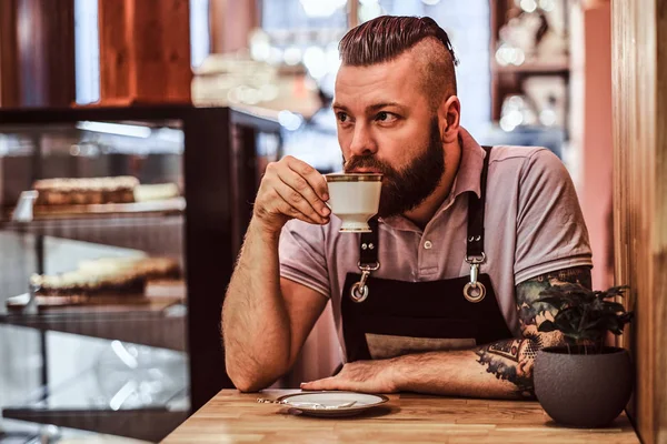 Guapo barista usando delantal bebiendo café durante el descanso del almuerzo sentado en una mesa en la cafetería —  Fotos de Stock