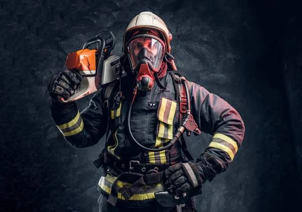 Firefighter wearing full protective equipment posing with a chainsaw on his shoulder. Studio photo against a dark textured wall — Stock Photo, Image