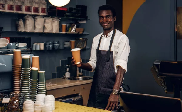 Barista africano sosteniendo una taza con café mientras está de pie detrás de un mostrador en una cafetería y mirando una cámara — Foto de Stock