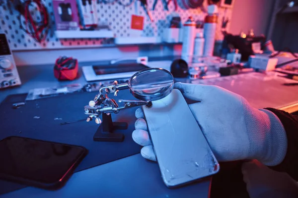 Técnico eletrônico detém um smartphone moderno com um corpo quebrado, examina cuidadosamente os danos usando uma lupa. Iluminação com luzes vermelhas e azuis — Fotografia de Stock