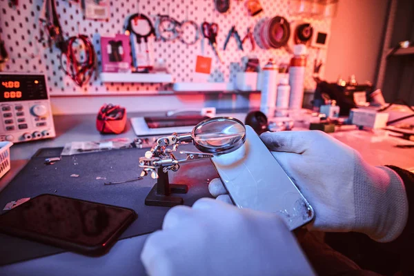 Técnico eletrônico detém um smartphone moderno com um corpo quebrado, examina cuidadosamente os danos usando uma lupa. Iluminação com luzes vermelhas e azuis — Fotografia de Stock