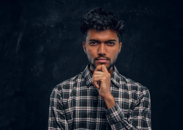 Bonito indiano cara em camisa xadrez olha para a câmera com um olhar piercing e segura a mão no queixo . — Fotografia de Stock