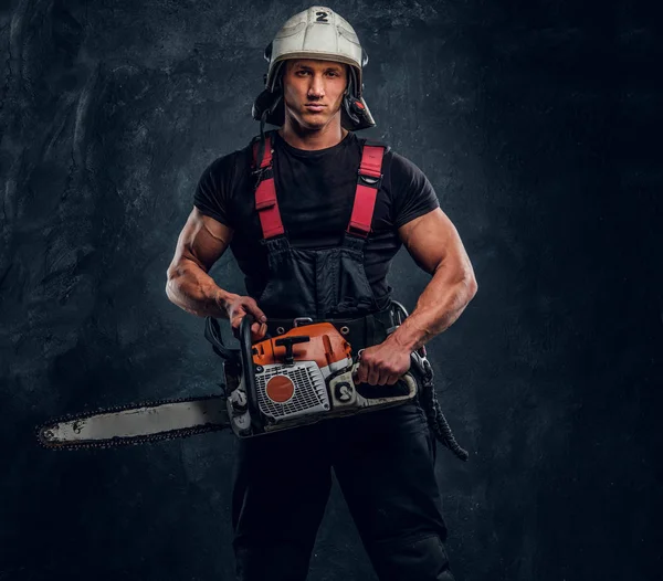 Young lumberjack wearing protective clothes posing with a chainsaw in a dark studio — Stock Photo, Image