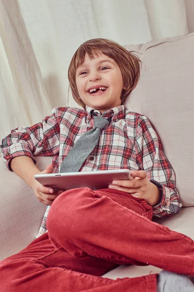 Glada skolpojke klädd i rutig skjorta med slips avkopplande på en soffa med en Tablet PC-dator hemma — Stockfoto