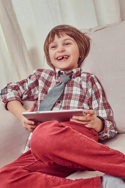 Veselý školák nosí kostkovanou košili s kravatou relaxační na gauči s tablet počítač doma — Stock fotografie