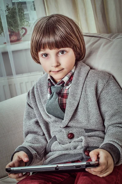 Ung skolpojke eleganta kläder håller en digital surfplatta medan du sitter på en soffa hemma. — Stockfoto