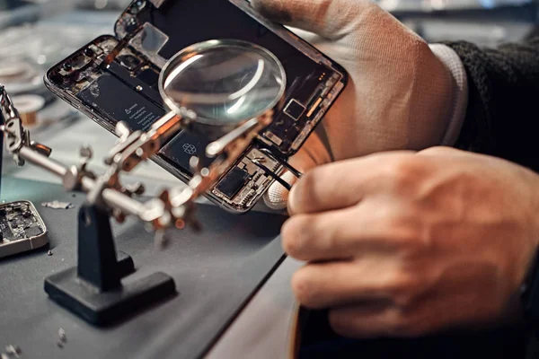 Reparador utiliza lupa y pinzas para reparar el teléfono inteligente dañado . —  Fotos de Stock