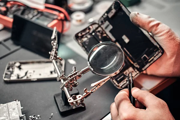 Reparador utiliza lupa y pinzas para reparar el teléfono inteligente dañado . — Foto de Stock