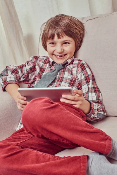 Веселый школьник в клетчатой рубашке с галстуком расслабляется на диване с планшетным компьютером дома — стоковое фото