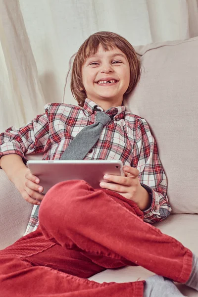 Wesoły uczniak kraciaste koszule na sobie krawat relaksujący na kanapie w domu na komputerze typu tablet — Zdjęcie stockowe