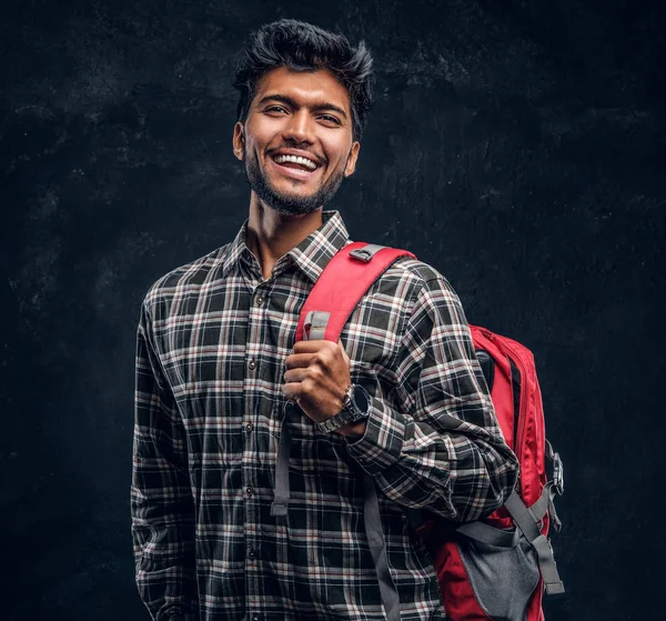 Ritratto di un bellissimo studente indiano con uno zaino che indossa una camicia a quadri, sorride e guarda una macchina fotografica . — Foto Stock