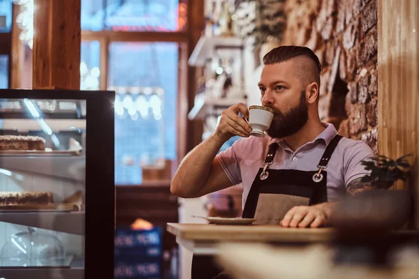 Guapo barista usando delantal bebiendo café durante el descanso del almuerzo sentado en una mesa en la cafetería — Foto de Stock